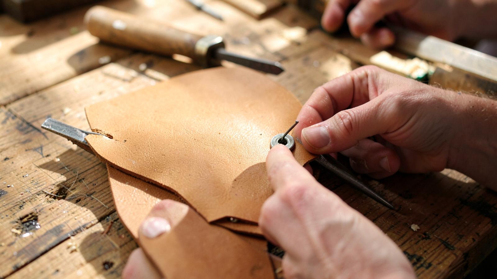 Craftsperson installing a prong magnetic snap into a tan leather bag flap with an awl and mallet on a workbench.