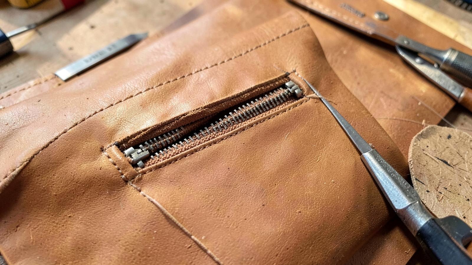 Zippered welt pocket set into a tan leather bag panel with a metal zipper and stitching tools nearby.